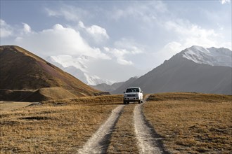 Off-road vehicle on a track between yellow meadows, glaciated high mountain peaks in the