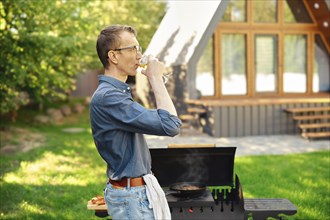 Middle-aged man drinks wine while standing in profile at the grill in the backyard of a log cabin