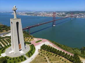 A tall statue with outstretched arms stands on a viewpoint near a bridge on the water, with a city