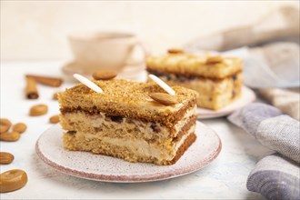 Honey cake with milk cream, caramel, almonds and a cup of coffee on a white concrete background and