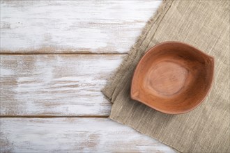 Empty clay brown bowl on white wooden background and linen textile. Top view, copy space, flat lay