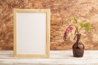 Wooden frame with pink dicentra, broken heart in ceramic vase on brown concrete background. side