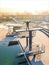 A person stretches his arms on a diving platform in an empty swimming pool in the sunlight, ENCW