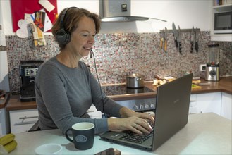 Woman, mid-50s, works from home, with laptop and communicates with colleagues via headset, home