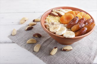 A plate with muesli, banana, dried apricots, dates, Brazil nuts on a white wooden background. close