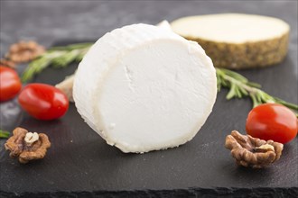 White goat cheese and various types of cheese with rosemary and tomatoes on black slate board on a