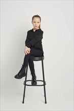 Young girl dressed in a stylish black outfit, sits casually on a high stool against a white