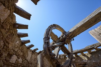 Abandoned building with an old wooden wheel and dilapidated stone walls under a clear blue sky,