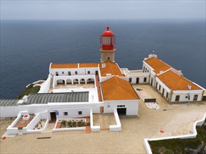 Close-up of a red lighthouse with several white buildings and a courtyard surrounded by the ocean,