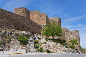 Historic castle with massive stone walls and towers, surrounded by trees and rocks under a clear