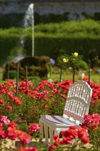 White baroque bench stands in a blooming garden full of red roses with a bubbling fountain in the