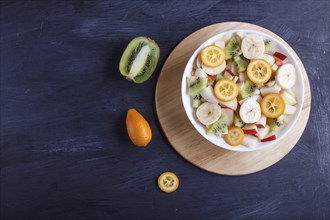 Vegetarian salad of bananas, apples, pears, kumquats and kiwi on black woodem background, top view,