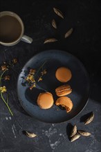 Orange macarons or macaroons cakes with cup of coffee on a black concrete background. Hard light,