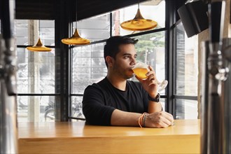 Man in black shirt sipping a glass of beer sitting at the bar in a modern bar with large windows.