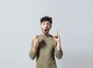 Amazed young man holding toothbrush pointing up. Guy with surprised gesture holding toothbrush