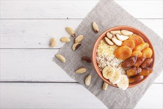 A plate with muesli, banana, dried apricots, dates, Brazil nuts on a white wooden background. top