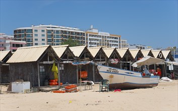 Beach houses and a painted boat on the beach with modern buildings in the background under a clear