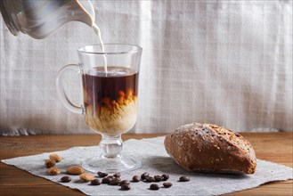 Glass cup of coffee with cream poured over and bun on a wooden background and linen textile. close