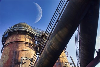 Rusted industrial plant in front of a clear blue sky with visible moon in the background,