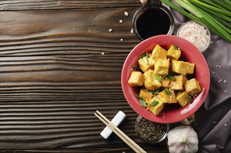 Flat lay view at stir fried tofu cubes with chives in clay dish on wooden kitchen table with napkin