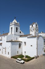 White church with two towers and clock on a paved square under a blue sky, Church of Santa Maria do