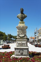 Stone bust on a pedestal with flowerbed in the foreground and clear sky in the background, monument