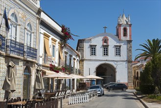 Street view with cafés, a white and red church and lush vegetation under a bright blue sky, house