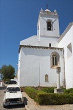 Church building with white tower and parked car in a narrow cobblestone alley, Santa Maria do