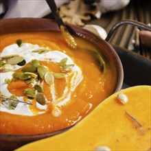 Clay dish with homemade rustic pumpkin soup with seeds on wooden table with bread and greens aside