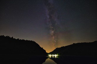 The Milky Way stretches across the night sky over a calm lake, framed by dark trees, Alp Stausee,