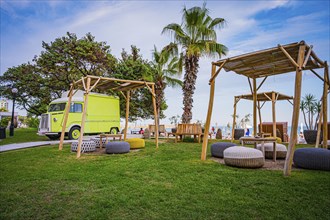 A yellow motorhome next to wooden canopies and palm trees on a green meadow near the sea, Golden