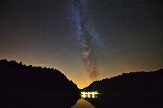 The Milky Way shines in the night sky above a quiet lake surrounded by dark trees, Alp Stausee,