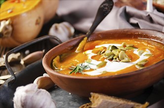 Clay dish with homemade rustic pumpkin soup with seeds on wooden table with bread and greens aside