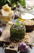 Glass jar with genovese pesto sauce on cutting board with basil leaves, pine nuts, parmesan cheese,