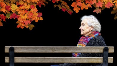 Elderly woman sitting on a bench under autumnal coloured leaves in front of a black background,