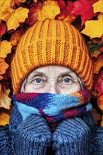 Elderly woman looks through colourful autumn leaves, warmly wrapped up with scarf and hat,