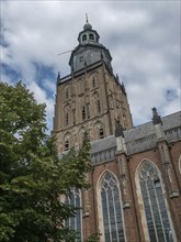 High church tower with gothic architecture, partly hidden by trees, under a cloudy blue sky,