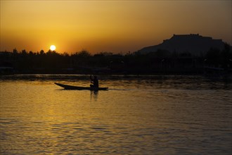 The silhouette of a boat glides along the surface of Dal Lake during sunset with Srinagar Fort in