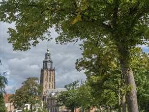 Tall historic church tower surrounded by lush trees and a green park under a blue, cloudy sky,