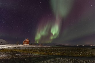 Green and violet northern lights over a red house on the coast, aurora borealis, winter, snow,