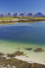 A calm, clear fjord with seaweed, white sandy beach and mountains in the background under a bright