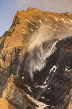 The spray of a waterfall sprays upwards, strong wind, mountains, morning light, East Iceland,