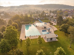 A large swimming pool surrounded by green areas and trees, several buildings, morning atmosphere