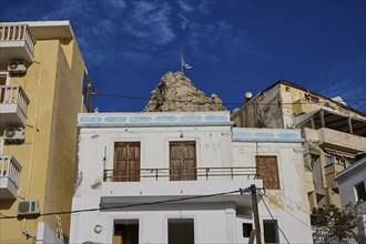 White and yellow buildings in a coastal town in front of rocks with the Greek flag under a clear