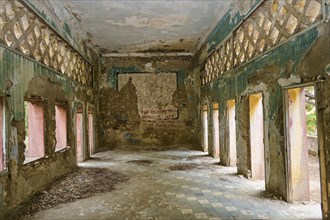 Abandoned room with large windows and peeling paint on walls, partially preserved decorations and