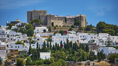 Castle on a wooded hill above white houses and cypresses, under a bright blue sky and warm