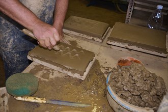 Worker's hand carving patterns in clay bricks, surrounded by clay and tools, Patmos Cotto