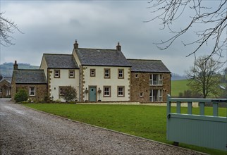 View of an older rendered house with a stone outbuilding behind a lawn and in front of a gravelled