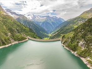 A reservoir with a dam, surrounded by mountains and green slopes, under a cloudy sky, Klein Tibet,