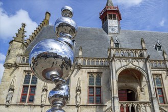 Market square reflected in shiny modern artwork in front of the Brabantine Gothic town hall in the